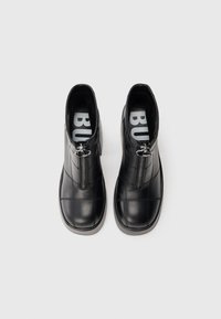 Botas pretas de cano curto em couro liso, com fecho lateral e padrão de costura cruzada. Biqueira arredondada e sola plana com ligeira elevação.