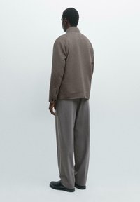 Brown wool blend jacket with a high collar, side pockets, and buttoned cuffs, paired with light grey wide-leg trousers and black shoes.