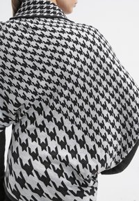 Czarnobiały płaszcz w wzór pepitki wykonany z miękkiego materiału; posiada szeroki kołnierz i opada na jedno ramię, eksponując swoją fakturę.