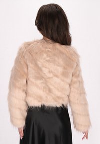 Faux fur cropped jack in lichtbeige met lange mouwen; zachte textuur met een licht golvend patroon; gedragen over een zwarte satijnen rok.