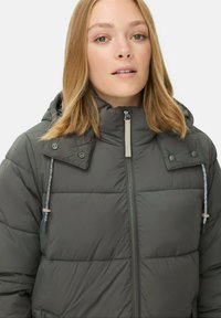 Mørkegrøn pufferjakke med høj krave, lynlåslukning og trykknapper. Har snoretræk ved hætten og quiltet tekstur.