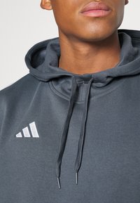 Hoodie gris foncé avec une texture douce, doté d'un cordon de serrage, d'une poche kangourou et d'un logo Adidas blanc réfléchissant.