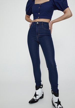Mujer con vaqueros ajustados de talle alto en azul oscuro, una camiseta cropped azul marino con mangas abullonadas y botines negros y blancos con estampado de estrellas.