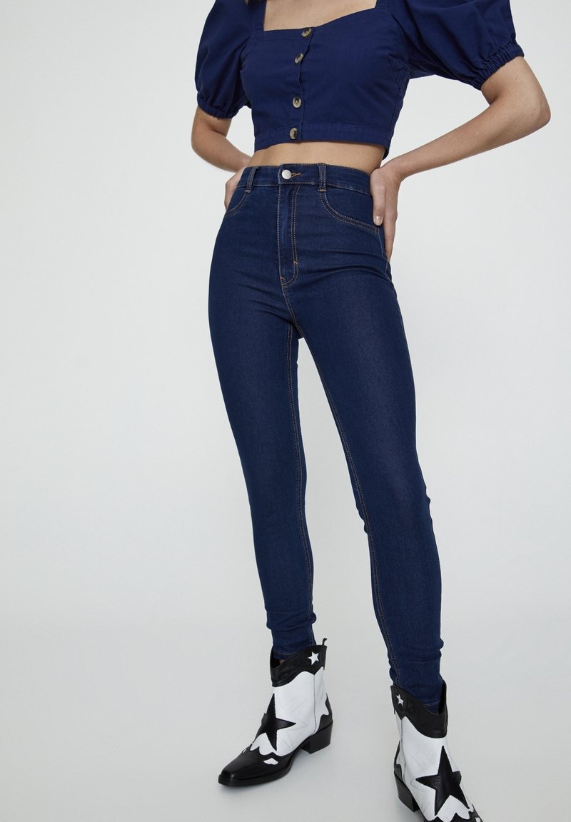 Femme portant un jean skinny taille haute bleu foncé, un top court bleu marine à manches bouffantes et des bottines noires et blanches à motif étoilé.