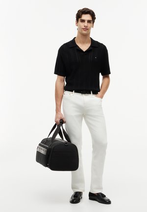 Young man wearing a black short-sleeve shirt and white pants, holding a black duffel bag, standing against a white background.