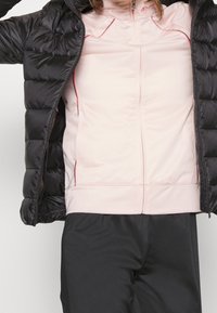 Person med en svart vadderad jacka över en blekrosa zip-up träningsskjorta och svarta byxor, armarna utsträckta ur jackans ärmar.