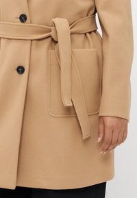 Kamel-farget, ull-blandingsfrakk med spiss krage, svarte knapper, belte i midjen og en lomme foran. Myk tekstur og strukturert design.