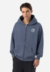 Siva zip-up dukserica izrađena od mekanog materijala. Ima veliki prednji džep, kapuljaču i logo na grudima. Labav kroj s spuštenim ramenima.