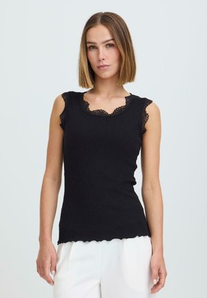 Ung kvinne med rett, skulderlangt hår, iført en tettsittende svart ermeløs topp med blonderkant og hvite bukser mot en ensfarget bakgrunn.