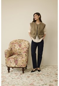 Olivengrøn ruskindsvest med to forlommer, lagdelt over en cremefarvet bluse, kombineret med mørke denim bukser og sorte spidse flade sko. Blomsterbetrukket lænestol i baggrunden.