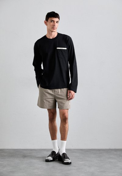 Svart langermet skjorte med brystlomme, kombinert med beige rutete shorts. Modell som har på seg hvite sokker og svarte sko. Glatt stofftekstur.