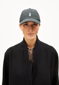 Dark green baseball cap with an adjustable strap, featuring a white embroidered ribbon logo on the front. Worn with a black high-collared coat.