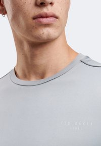 Κοντινό πλάνο στο κάτω μέρος του προσώπου και τον λαιμό ενός ατόμου που φορά ανοιχτογκρι μπλουζάκι Ted Baker Sport με σκουλαρίκι κρίκο στο αυτί.