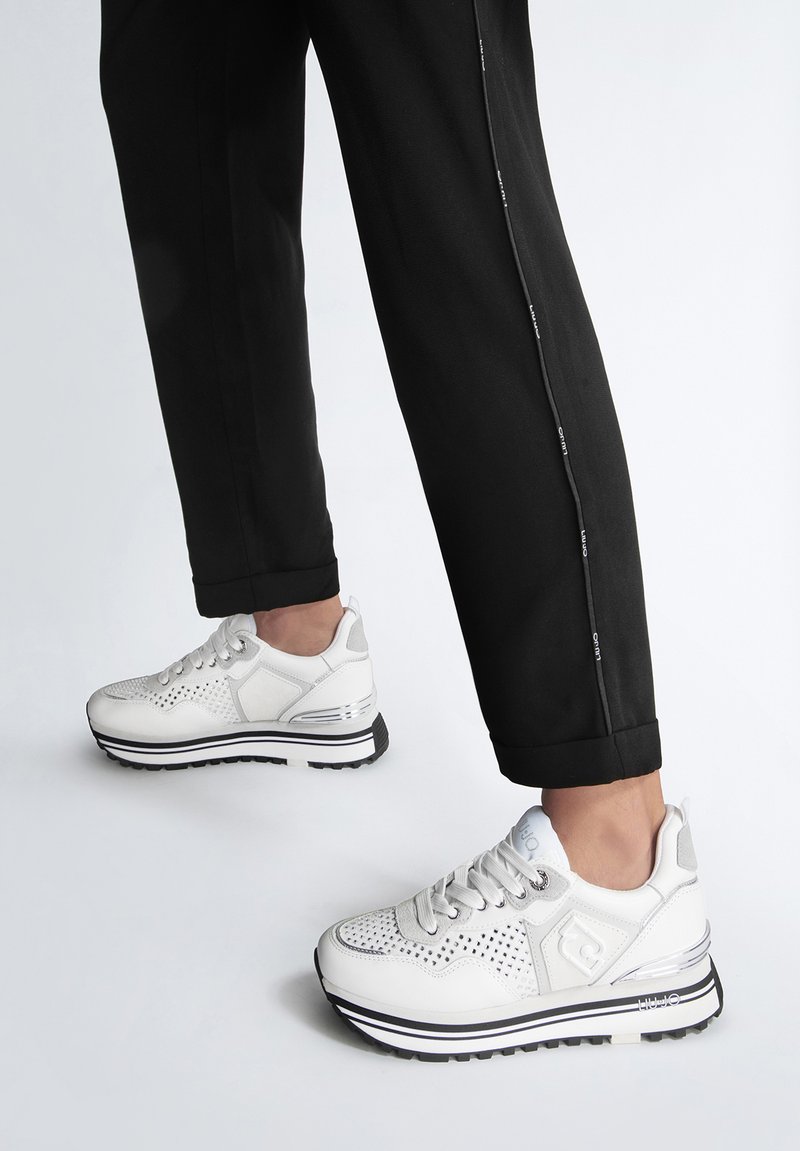 Sneakers atletici bianchi con una tomaia in rete e pelle, dettagli perforati e una suola a strisce bianco e nere. Abbinati a pantaloni neri.