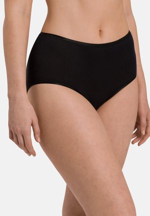 Vrouw die effen zwarte hoogwater slips draagt, deels zichtbaar vanaf de taille tot aan de bovenkant van de dijen, staand tegen een witte achtergrond.