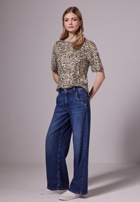 Top s krátkým rukávem v leopardím vzoru v béžové a černé barvě, kombinovaný s modrými džíny s širokými nohavicemi. Celkový look doplňují bílé tenisky.