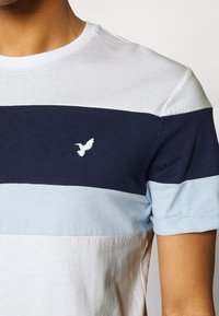 T-skjorte med kort erm, med hvit, marineblå og lyseblå stripet design. Inkluderer en brodert fuglelogo på venstre brystområde. Myk tekstur.