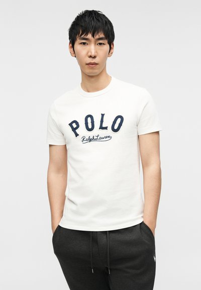 Λευκό βαμβακερό μπλουζάκι με το λογότυπο «POLO Ralph Lauren» σε σκούρο μπλε γράμματα. Το μοντέλο φοράει σκούρο γκρι παντελόνι φόρμας με κορδόνι στη μέση. Απλός σχεδιασμός.