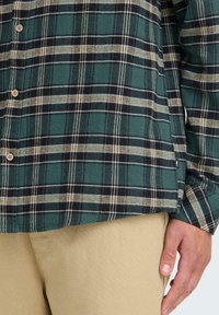 Grøn og sort plaid flannel skjorte med knapper, vist sammen med beige bukser. Skjorten har en blød tekstur og kontrasterende lyse striber.