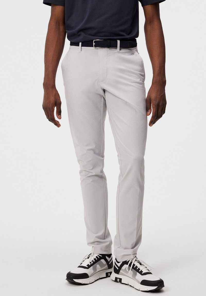 Světle šedé slim-fit kalhoty vyrobené z hladké tkaniny, s klasickým designem, černým tkaným páskem a bílými sportovními botami.