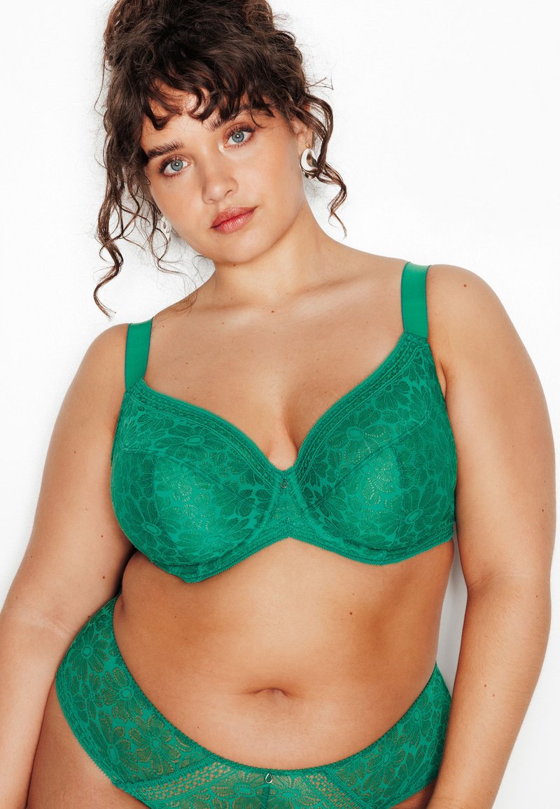 Femme aux cheveux bouclés portant un soutien-gorge et une culotte en dentelle florale verte assortis, face à un fond blanc, regardant droit devant avec une expression neutre.