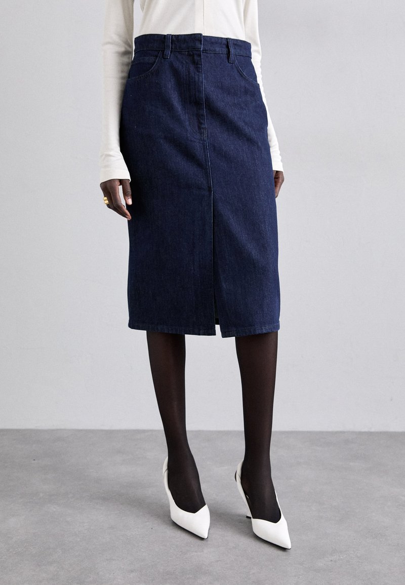 Donkerblauwe denim kokerrok met een split aan de voorkant en een klassieke vijf-pocketontwerp, gedragen met zwarte panty's en witte puntige hakken.