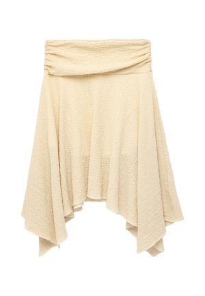Kvinde iført en hvid off-shoulder top, asymmetrisk beige nederdel og brune ruskindsstøvler til knæene, står mod en hvid baggrund.