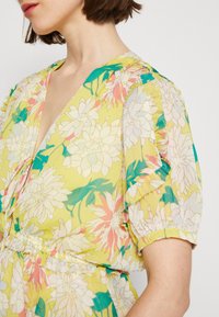 Blomklänning i gul tyg med rosa och gröna blommönster. V-ringning, puffärmar och rynkad midjedetalj. Lättviktig textur.