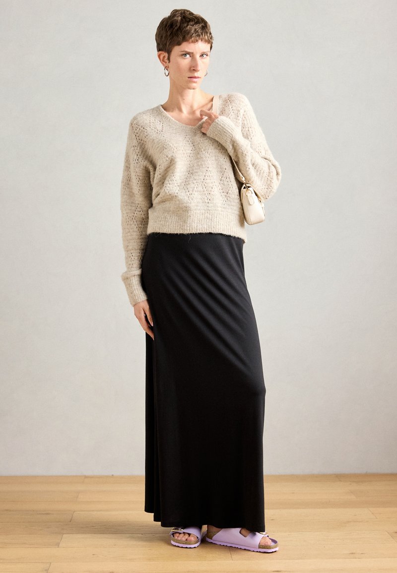 Person mit kurzen Haaren, die einen beigen Strickpullover, einen schwarzen Maxirock und lavendelfarbene Sandalen trägt und eine kleine beige Umhängetasche bei sich hat.