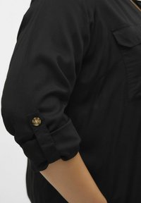 Sort skjorte med opsmøgede ærmer, der har en guldknap som accent. Blødt stof med en glat tekstur og en let glans. Minimalistisk design.