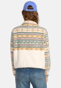 Chaqueta de forro polar con un diseño de patrón multicolor que presenta olas y formas geométricas, sección inferior blanca y cuello con solapa.