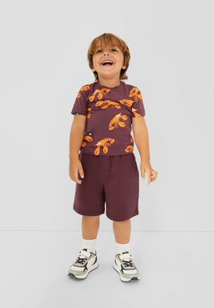 Jong kind lacht en staat, draagt een paarse shirt met oranje kreeftenprints, bijpassende korte broek, witte sokken en witte sneakers.