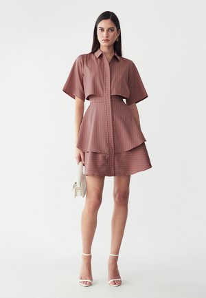 Mujer con vestido de dos piezas de rayas finas color malva, con mangas cortas, sosteniendo un pequeño bolso blanco y llevando tacones blancos con pulsera en el tobillo.