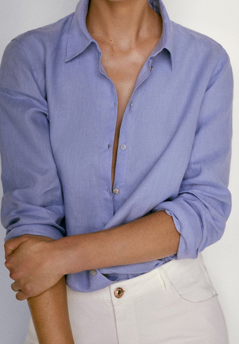 Camicia azzurra chiara con un taglio morbido, maniche arrotolate e una texture morbida. Abbinata a pantaloni bianchi con un bottone metallico.
