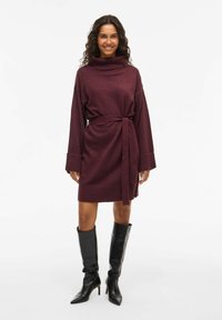 Robe en tricot bordeaux avec un col montant, des manches longues et une ceinture à nouer assortie ; accompagnée de bottes à talons noires. Texture lisse.