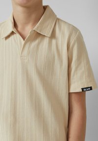 Beige polo shirt met een gestructureerd visgraatpatroon, een klassieke kraag en korte mouwen. Zwarte label met "SLVR" op de zoom.