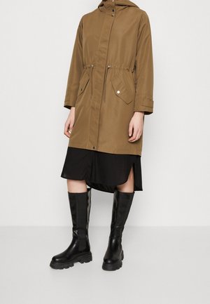 Brown hooded parka with drawstring waist, two front pockets, and button closure. Paired with a black dress and knee-high black rubber boots.