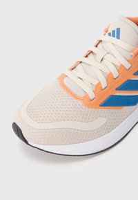 Primer plano de una zapatilla de correr beige con acentos naranjas, rayas azules, cordones blancos y parte superior de malla texturizada sobre fondo blanco.
