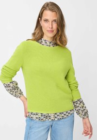 Maglione verde brillante lavorato a maglia con texture a coste, caratterizzato da maniche lunghe con motivo beige con onde scure, sovrapposto a jeans azzurri chiari.