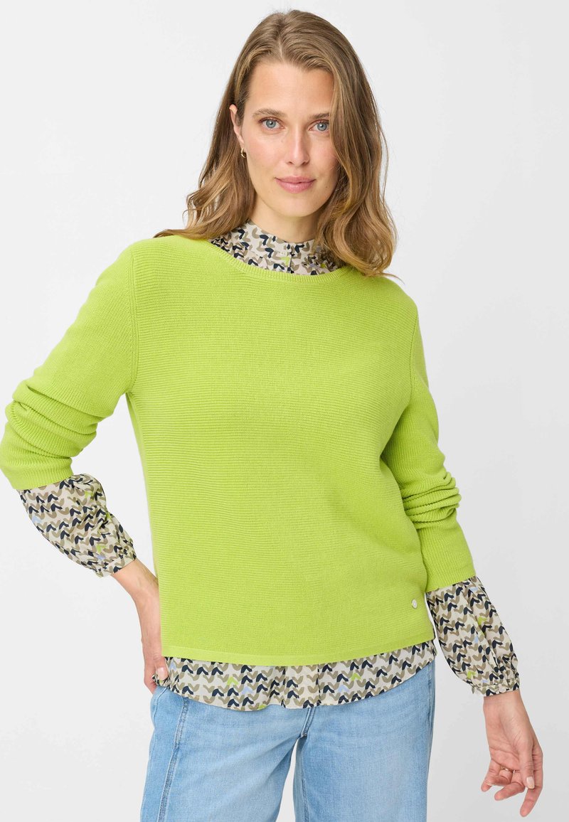 Maglione verde brillante lavorato a maglia con texture a coste, caratterizzato da maniche lunghe con motivo beige con onde scure, sovrapposto a jeans azzurri chiari.
