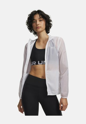 Mujer con sujetador deportivo negro con logo parcial, leggings negros y una chaqueta blanca translúcida con cremallera y capucha, de pie contra un fondo blanco.