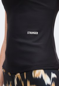 Μαύρο εφαρμοστό αθλητικό τοπ με τη λέξη «STRONGER» τυπωμένη σε άσπρο στο στήθος, σε συνδυασμό με παντελόνια με σχέδιο.
