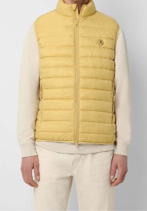 Mann trägt eine gelbe gesteppte ärmellose Daunenweste mit Frontreißverschluss über einem beigen Langarmpullover und beigen Hosen.