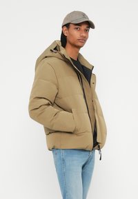 Polstret jakke i tan farve med hætte, lynlås-lukning og sidelommer. Båret over en sort skjorte, parret med lyseblå jeans.