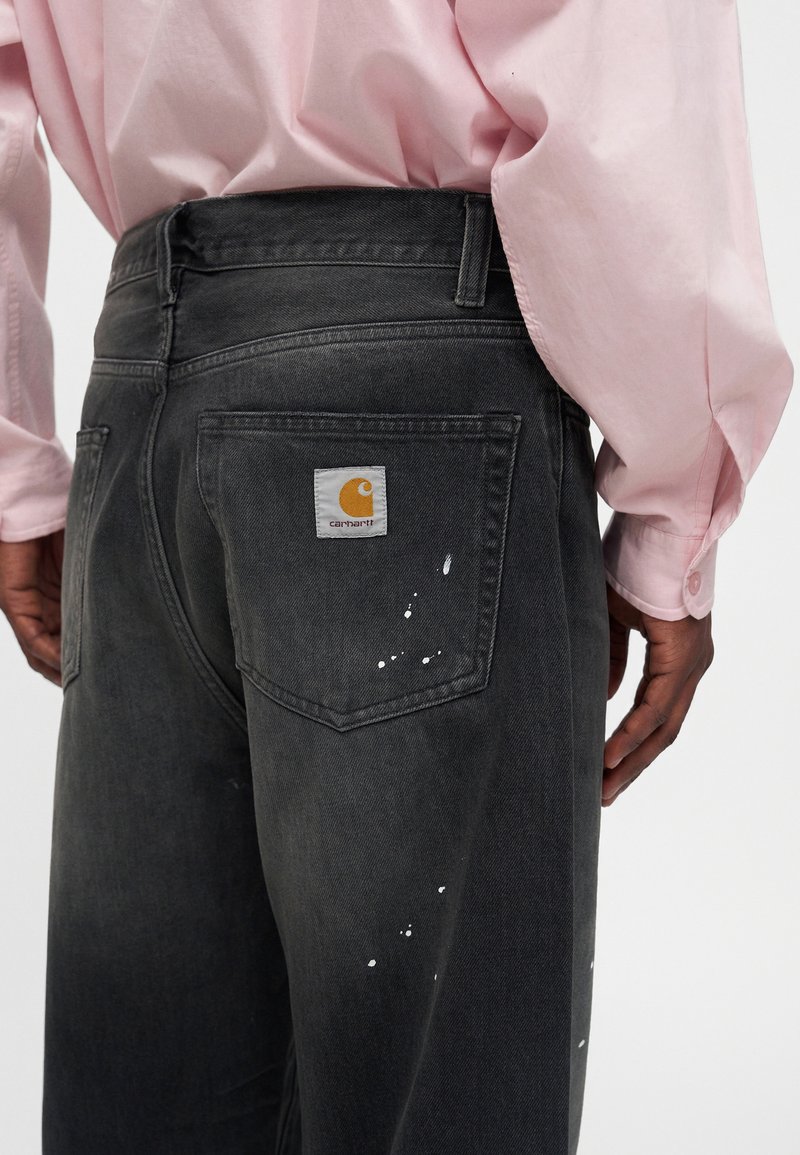 Person wearing black Carhartt jeans with white paint splatters and a pink long-sleeve shirt, shown from the back waist down.