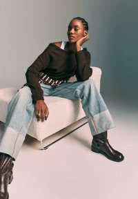 Bruine gebreide trui, lichtblauwe spijkerbroek met onafgebroken zoom, bruine leren enkellaarsjes met zilveren hardware, zittend op een witte gestructureerde bank.