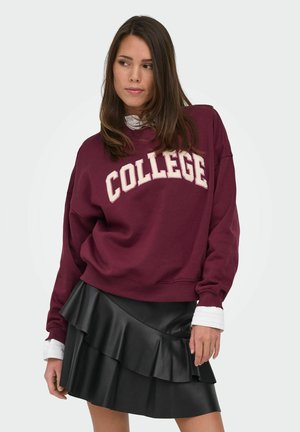 Jeune femme portant un sweat-shirt bordeaux avec le texte "COLLEGE" et une jupe en cuir noire superposée, regardant vers sa gauche.