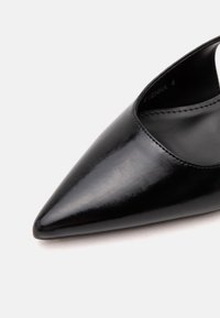 Gros plan d’une chaussure en cuir noire à bout pointu, surface lisse et brillante avec des coutures visibles le long du bord.