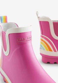 Stivali di gomma rosa con rifiniture bianche, caratterizzati da un motivo arcobaleno sul lato e un'etichetta multicolore sul retro. Label del marchio visibile.