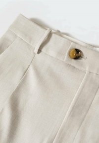 Gros plan de la taille d’un pantalon beige clair avec passants pour ceinture et fermeture par bouton rond marron sur fond blanc.